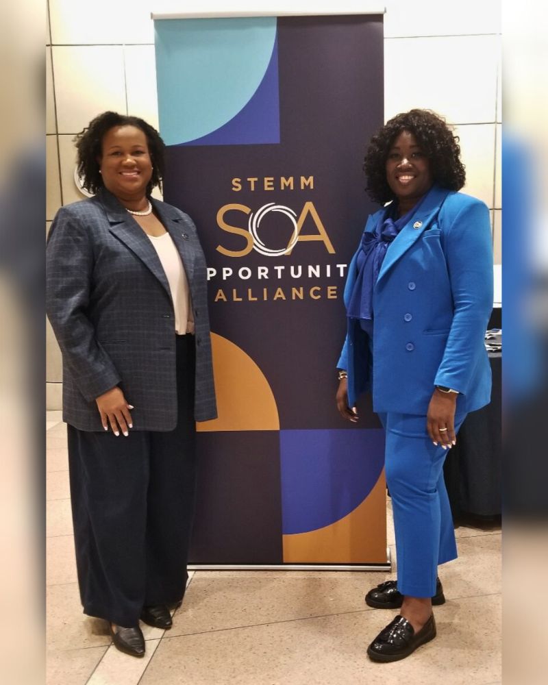 Two women in professional attire standing beside a banner that reads "STEMM opportunity alliance" in a technology conference hall.
