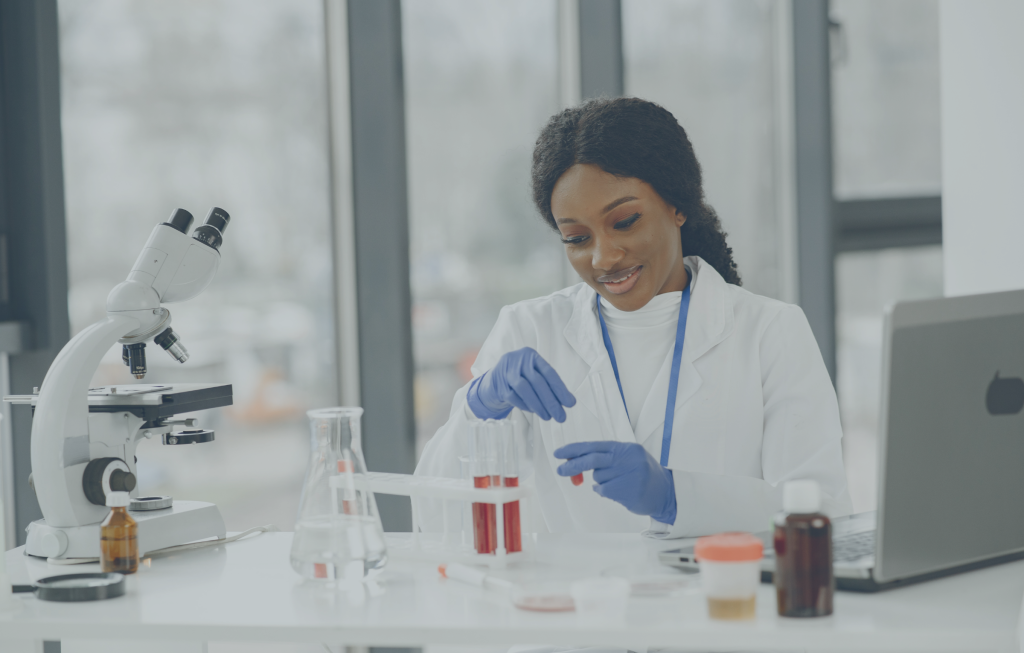 A scientist in a white lab coat and blue gloves conducts experiments with test tubes in a laboratory setting, surrounded by lab equipment, a microscope, and a laptop, all part of an innovative project supported by the Last Mile Fund.