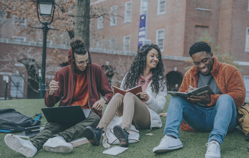 Three people sit on grass outside, smiling and looking at books and a laptop. A building and trees are visible in the background, embodying the spirit supported by initiatives like the Last Mile Fund.