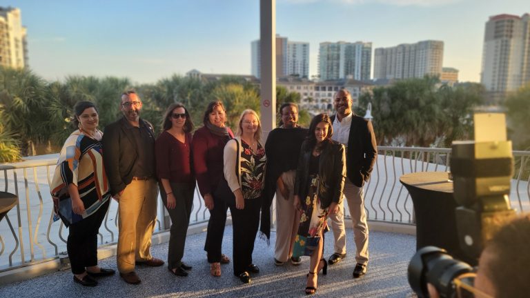 A group of people stands on a balcony with a cityscape and palm trees in the background, smiling at the camera.