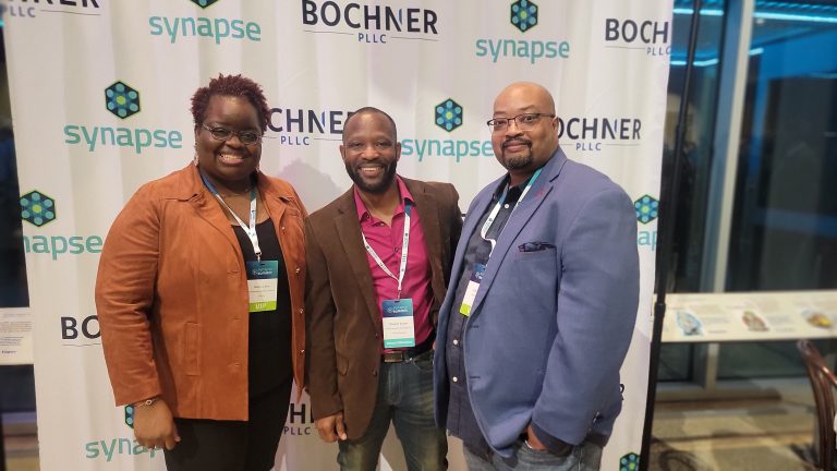 Three people smiling in front of a backdrop with "Bochner" and "Synapse" logos.