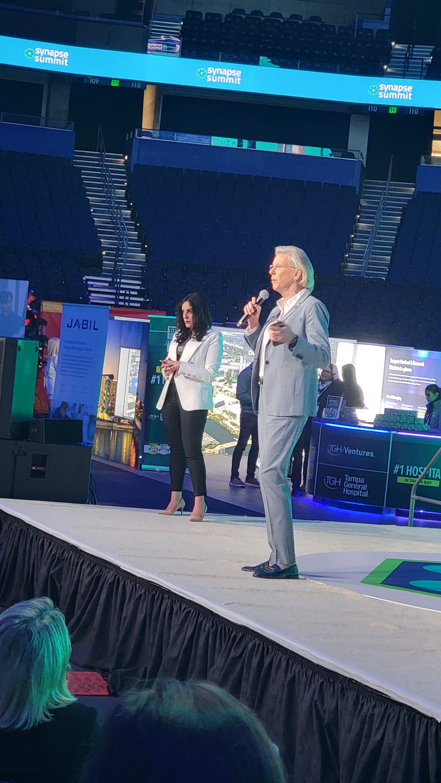 Two speakers on stage at the Synapse Summit, with a nearly empty audience seating area in the background.