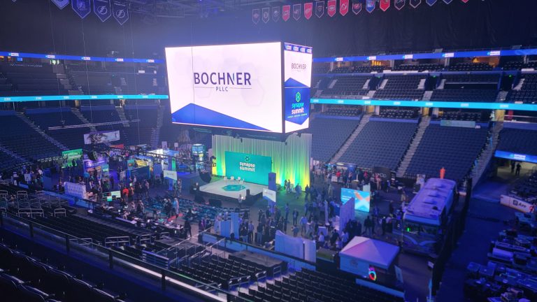 Indoor arena with a large screen displaying "Bochner PILC" above booths and attendees. The stadium seating is mostly empty.