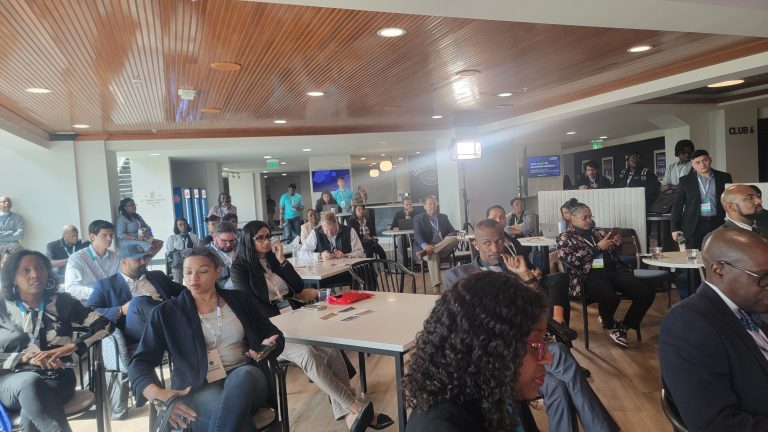 A group of people sitting and listening in a conference room with wooden ceiling and white walls. Some individuals are taking notes.