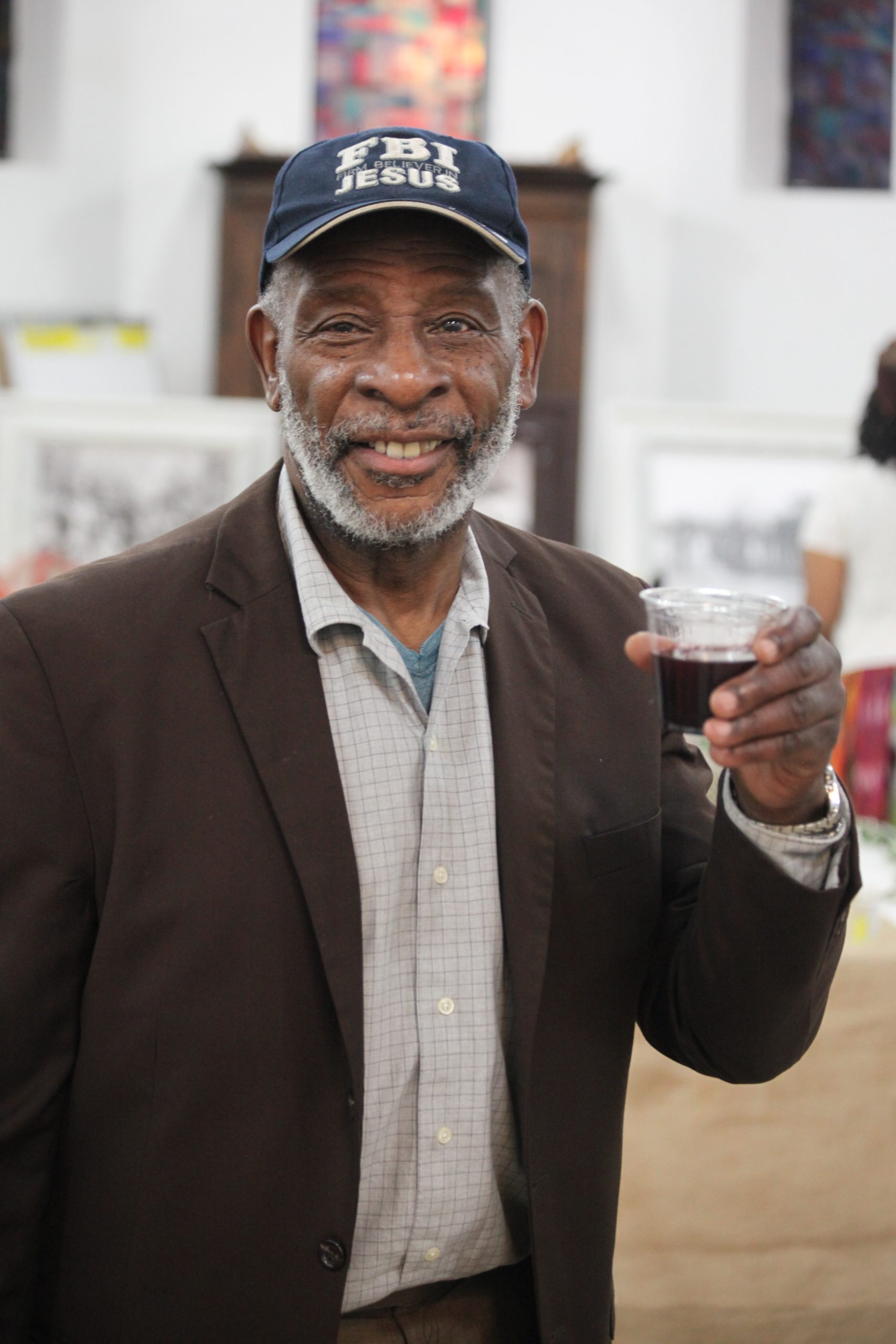 Man wearing a cap and holding a cup, smiling at the camera in an indoor setting.