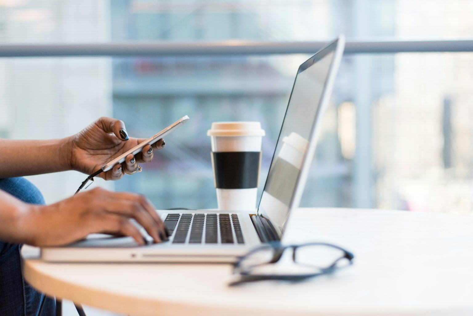 A person is at a table, adeptly managing an innovative digital pipeline. They're using a laptop and holding a smartphone, with a coffee cup and eyeglasses resting nearby.