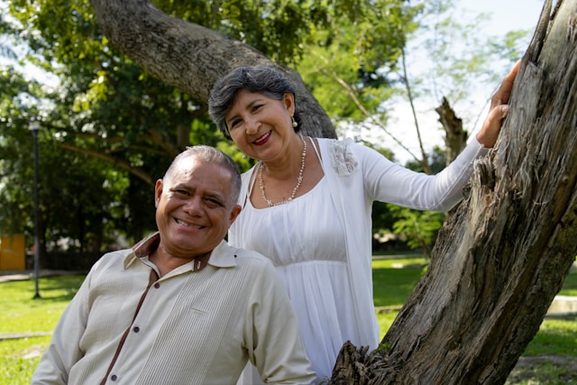 Older man and woman smiling outdoors, standing near a tree in a park on a sunny day, enjoying the benefits of the Pathways to Prosperity initiative.