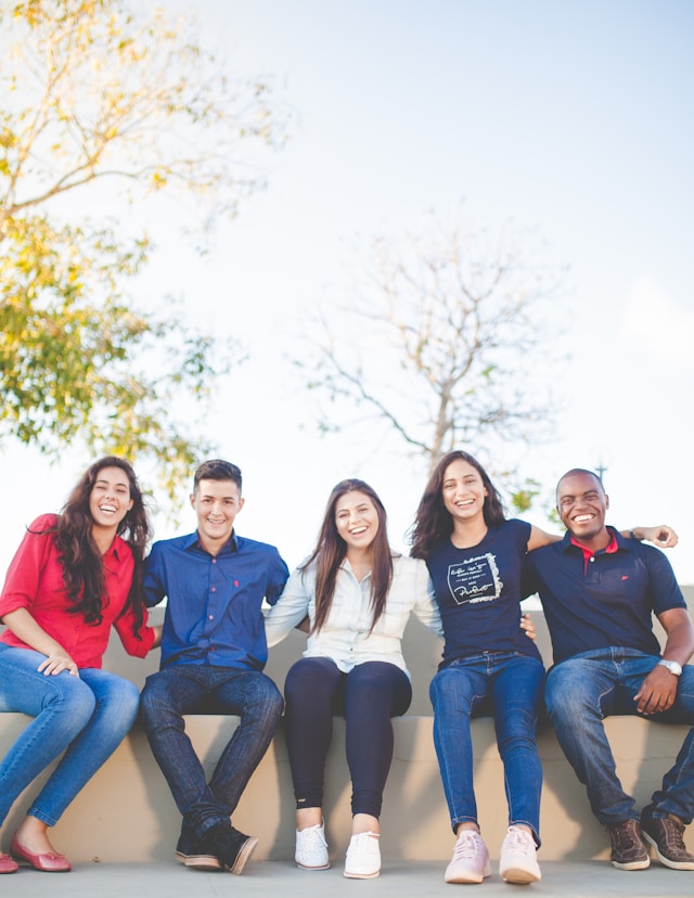 Five people sit outdoors on a low wall, smiling at the camera. Casually dressed, they enjoy a sunny day beneath leafy trees—a moment of connection inspired by the spirit of the Pathways to Prosperity Fund.